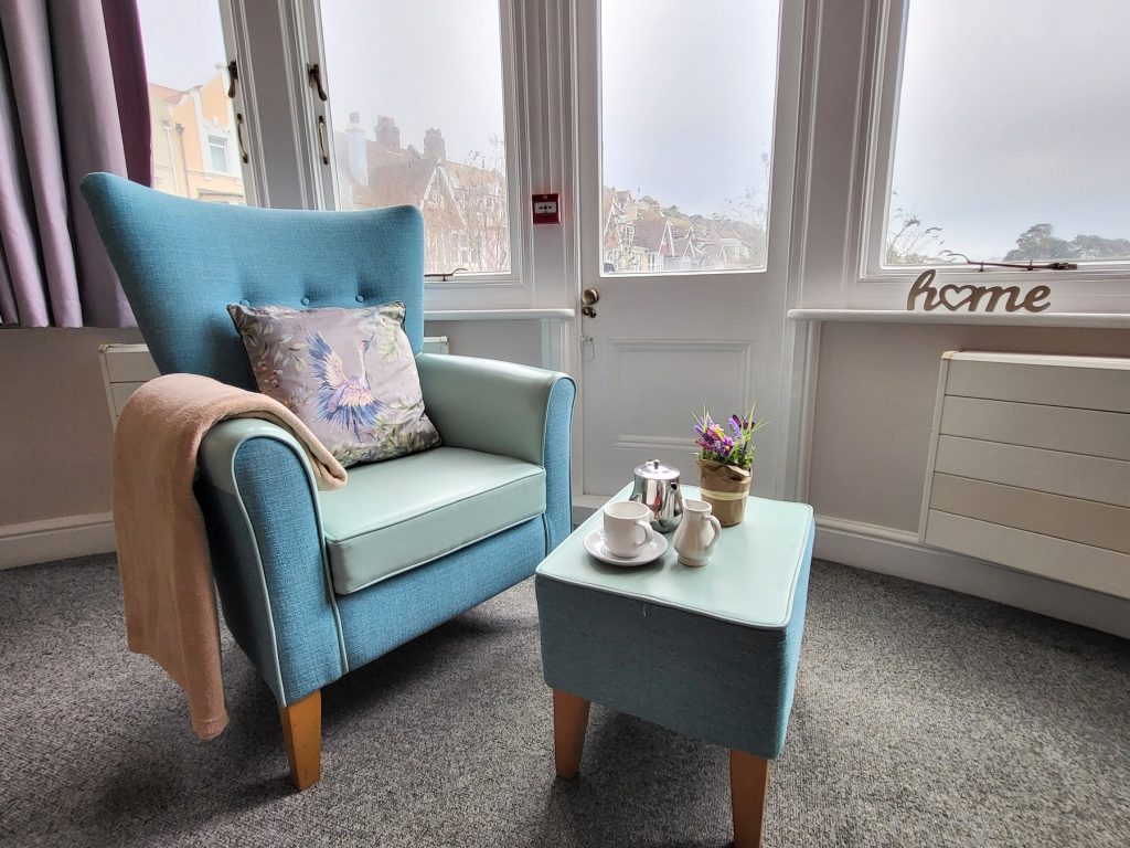 Grosvenor House care home lounge seating with teal armchair, side table and “home” sign