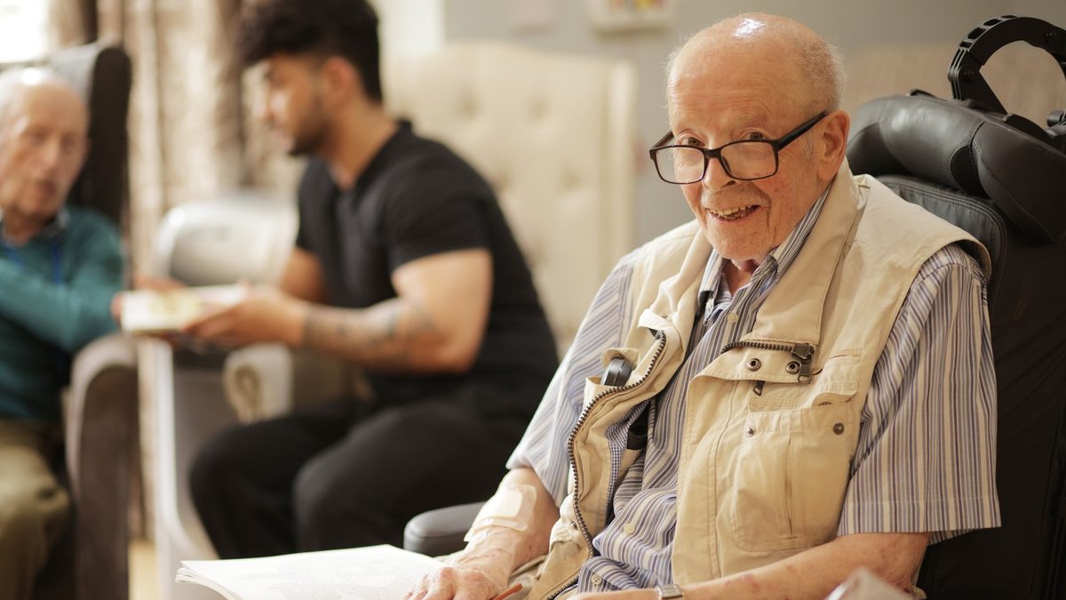 A smiling resident in glasses with a colouring book, looking at the camera, while a colleague chats with another resident behind him
